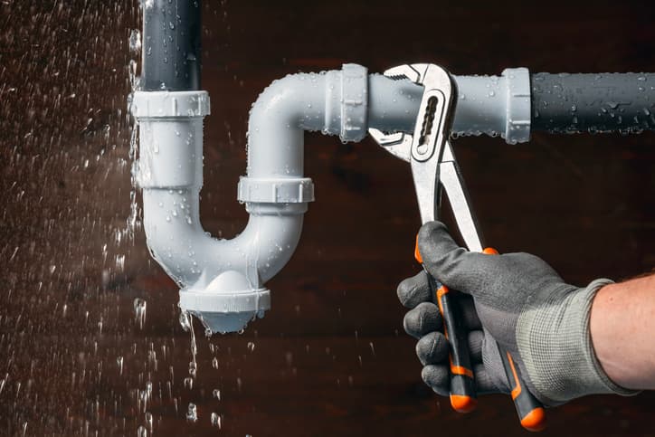 Person using pliers to repair a leaking pipe with water spraying from the connection